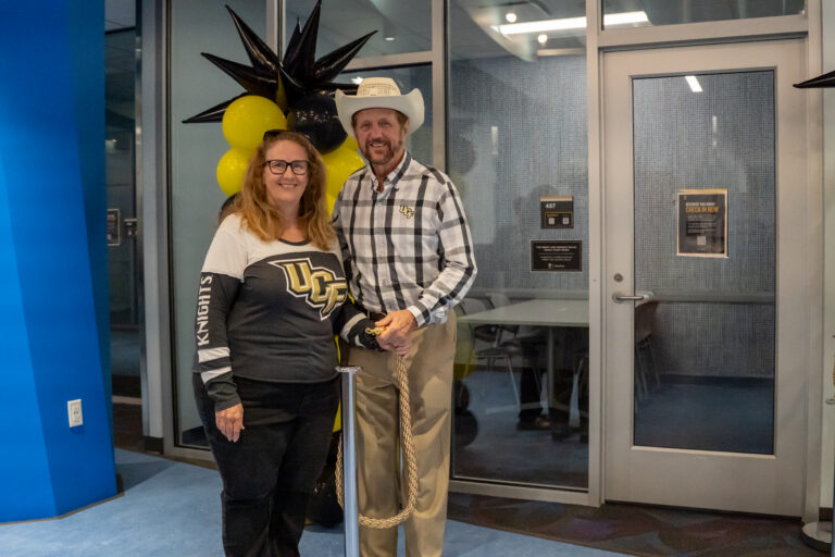 Smiling woman and man pose in front of glass doors with yellow and black balloons in the background at a lobby event.