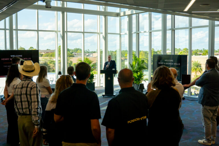 Speaker at a podium addressing a seated audience in a bright glass-walled room; screens say 'Welcome Henry & Georgia Willis'.