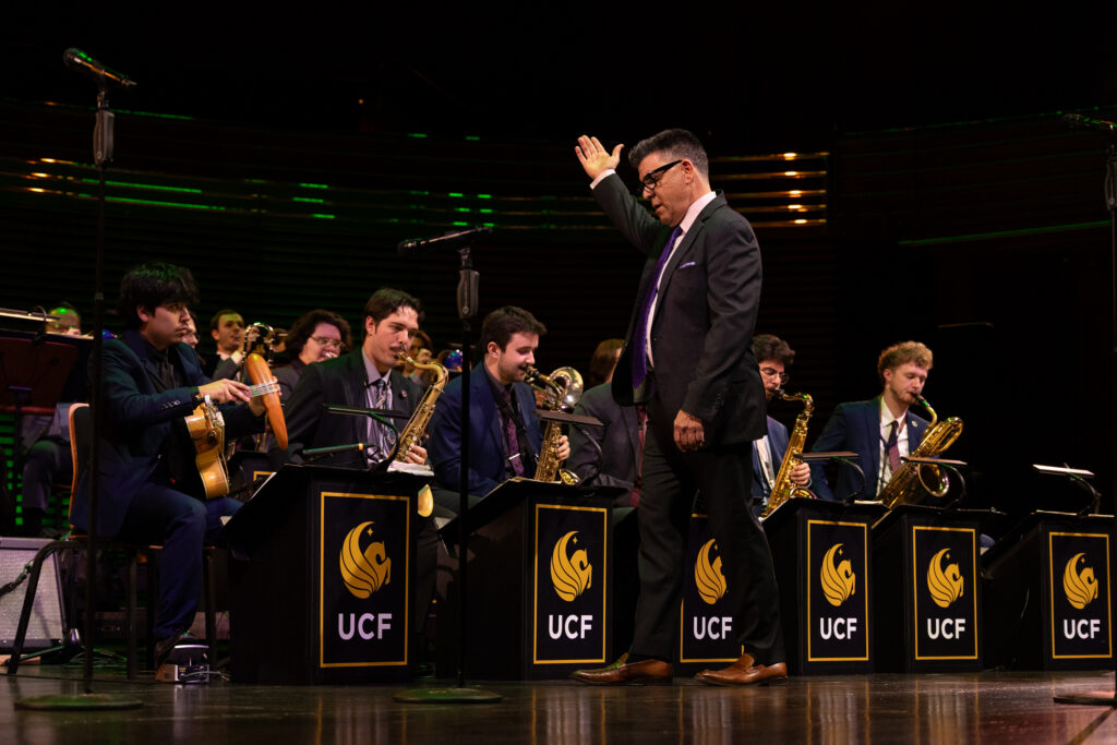 Conductor in a dark suit raises his hand while leading a university jazz band; musicians play saxophones and trumpets on stage with UCF stands behind them.
