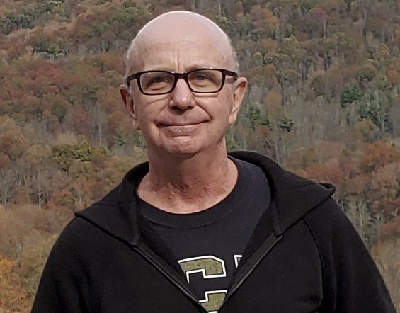 Smiling middle-aged man with glasses in a black zip-up hoodie outdoors, autumn trees in the background.